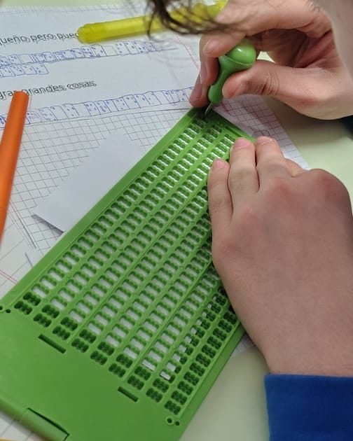 Manos de un alumno que escribe en braille con punzón sobre una pizarra verde