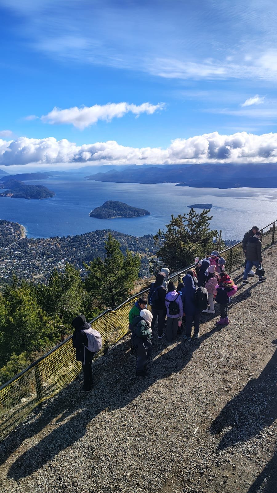Grupo de estudiantes y adultos en un mirador de montaña con baranda, frente al lago Nahuel Huapi y sus islas, bajo un cielo azul con nubes