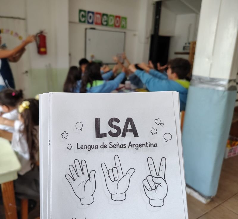 Cuadernillo «LSA – Lengua de Señas Argentina» en primer plano; detrás, el grupo practica señas con las manos en alto