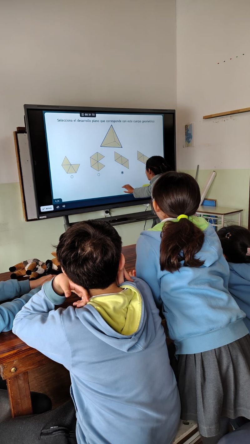 Alumnas y alumnos de espaldas frente a una pantalla inteligente del aula, mientras una compañera elige el desarrollo plano de una pirámide