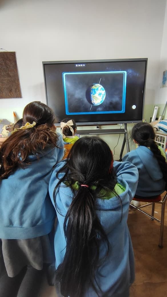Alumnas de espaldas, con el buzo celeste del uniforme, miran el planeta Tierra girando en una pantalla inteligente